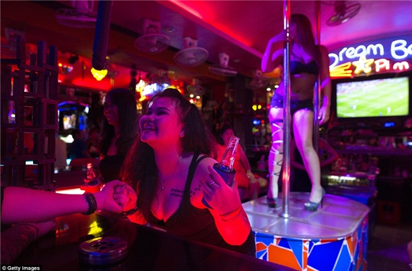 
Một cô gái bar đang lả lơi tiếp chuyện khách trong khi đằng sau là một đồng nghiệp đang biểu diễn múa cột.